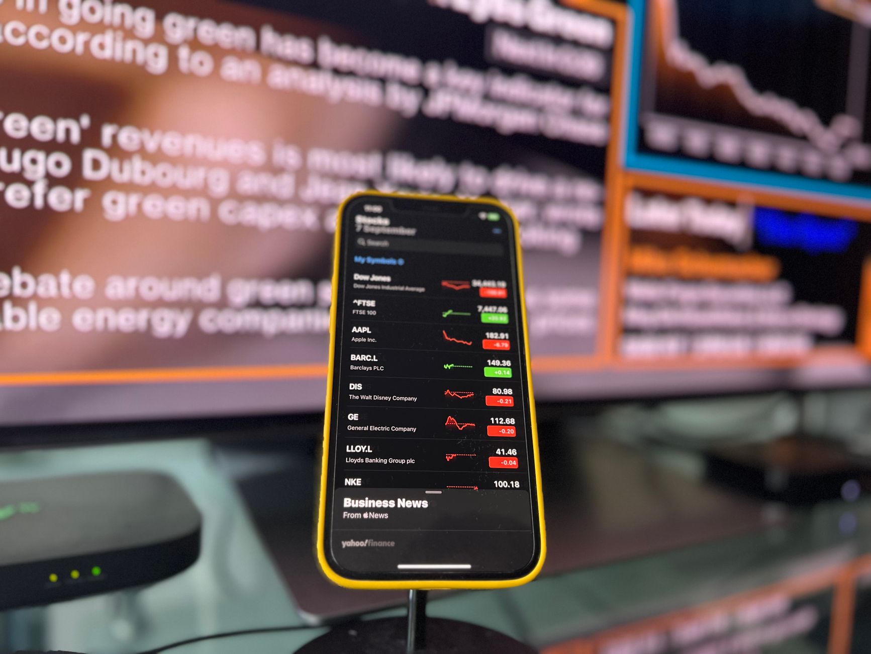 Celular com cotações diante de telas de notícias e gráficos do mercado financeiro
